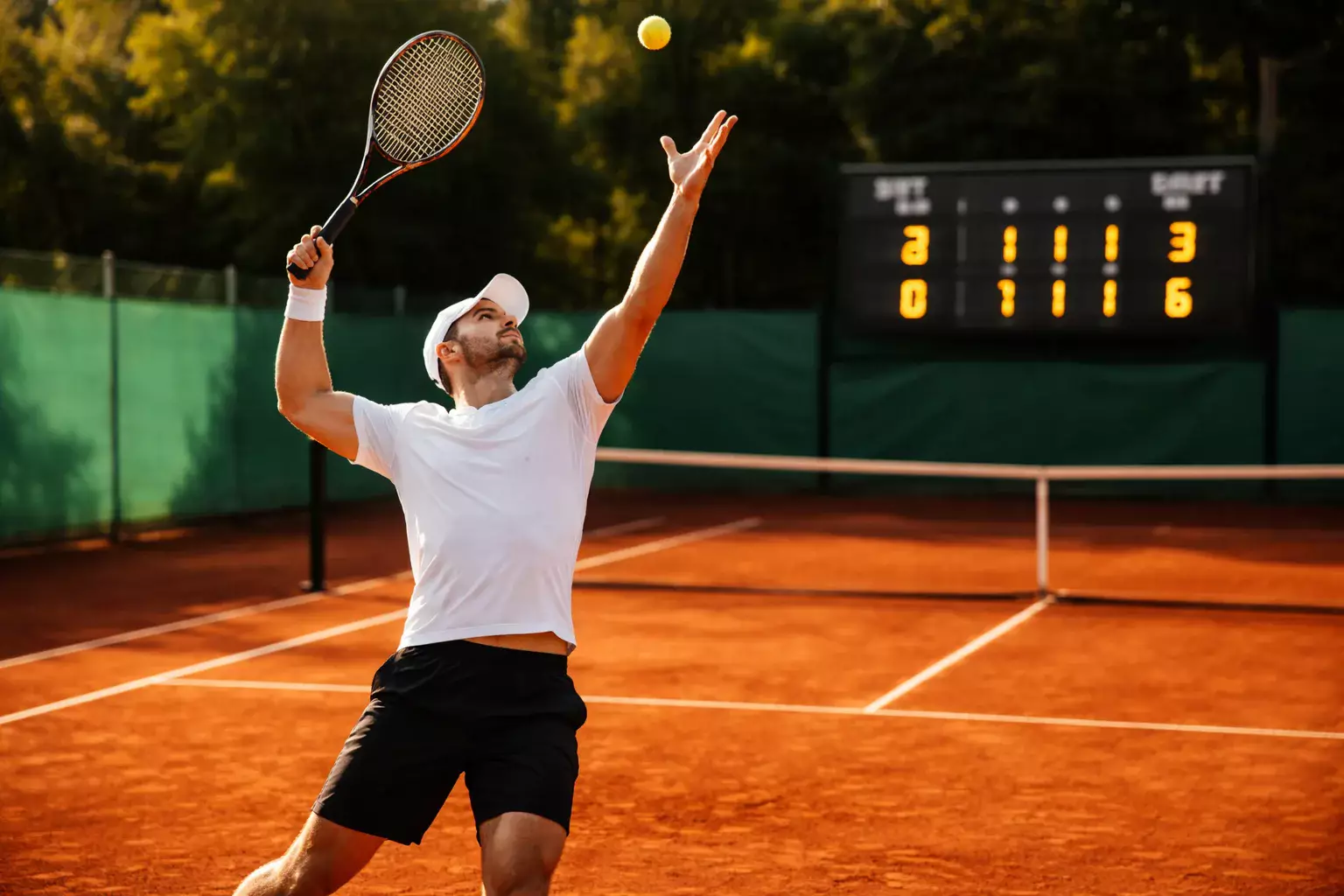 Tennisser serveert op een gravelcourt met het scorebord op de achtergrond